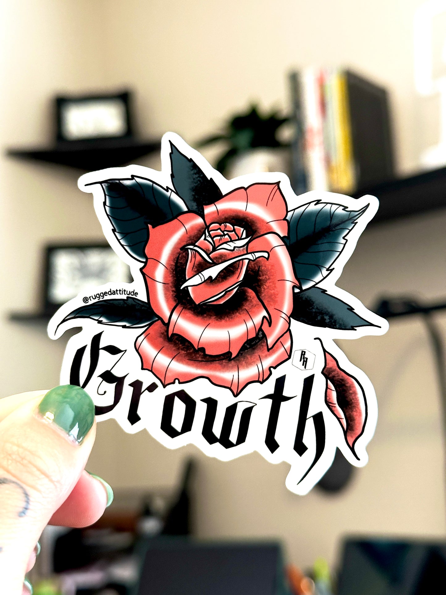 3x3 inch die cut sticker held in studio by artist, with a design of a rose with a fallen petal and gothic calligraphy word "growth". Branded with Rugged Attitude logo.