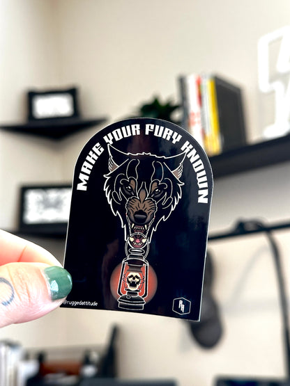 2x3 inch die cut sticker, held in studio by artist with the design of a wolf holding a lantern and gothic calligraphy words "make your fury known". Branded with Rugged Attitude logo.