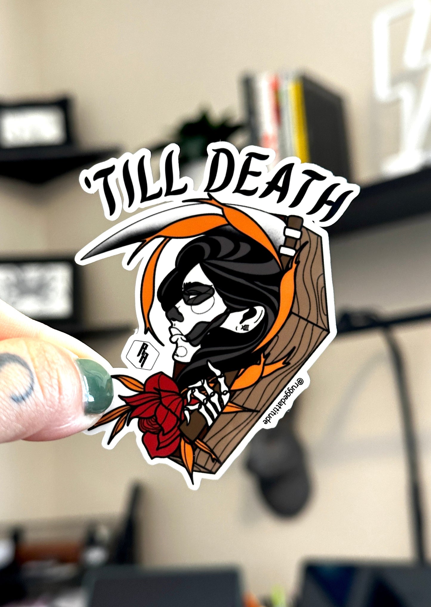 a 2.5 inch die cut sticker held in studio by artist, with a design of a female skull holding a scythe in a coffin with floral accent and words "till death". Branded with Rugged Attitude logo.